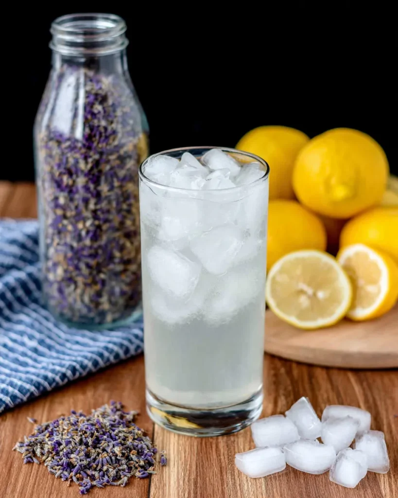 Top-down view of ingredients for Lavender Lemonade Cocktail, including a bottle of lavender-infused vodka, fresh lemons, dried lavender buds, a glass of lemonade, and ice cubes arranged on a rustic wooden table.