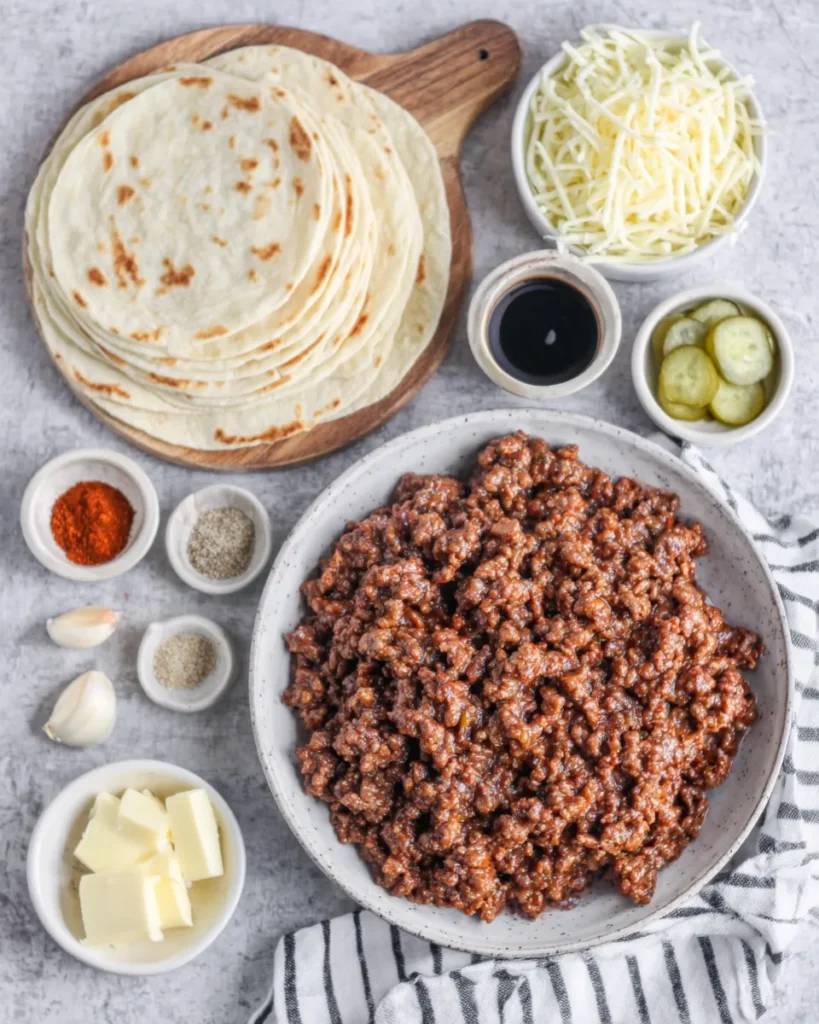 Separate plates and small bowls displaying ingredients for Easy Garlic Butter Beef Cheeseburger Rollups, including ground beef, shredded cheddar, shredded mozzarella, olive oil, flour tortillas, butter, garlic cloves, Worcestershire sauce, onion powder, garlic powder, smoked paprika, salt, black pepper, and sliced pickles.