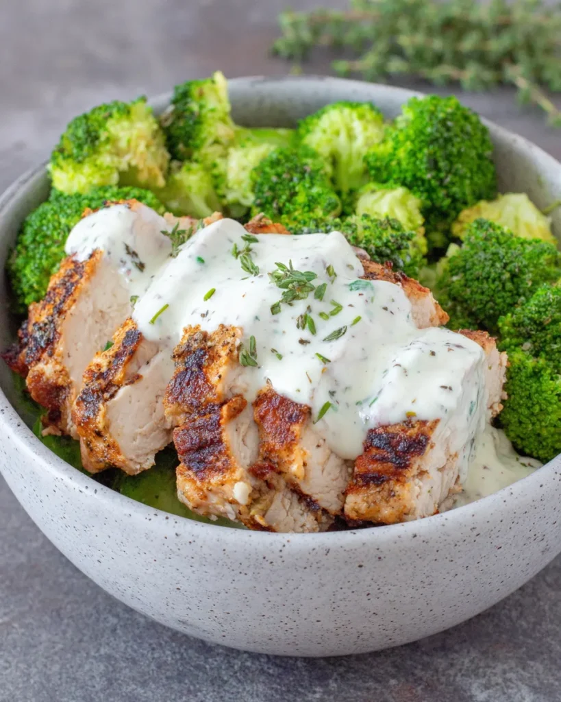 Grilled chicken and broccoli bowl drizzled with creamy garlic sauce, served in a white bowl with fresh parsley garnish, homemade dinner close-up.