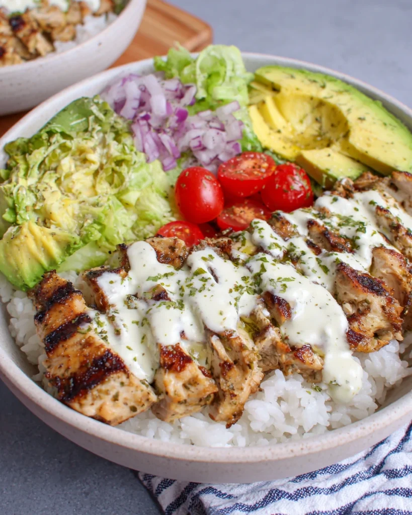 Close-up of a homemade grilled chicken rice bowl with sliced grilled chicken, fluffy white rice, avocado, cherry tomatoes, lettuce, and creamy herb sauce served in a white bowl on a marble kitchen counter with natural window light.