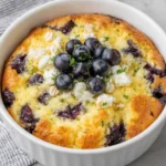 Close-up of a High Protein Blueberry Cottage Cheese Breakfast Bake in a white baking dish, featuring a creamy texture with fresh blueberries on top. Natural window light highlights the golden edges and homemade rustic appearance on a white marble kitchen counter.