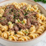 Close-up of homemade Slow Cooker Beef & Noodles served in a rustic bowl with tender shredded beef, egg noodles, and rich savory broth, photographed in natural kitchen lighting on a white marble countertop.