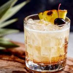 Close-up of a homemade Piña Colada Milk Punch served over ice in a vintage glass with pineapple garnish and maraschino cherry on a white marble kitchen counter, photographed in natural window light with a cozy homemade aesthetic.