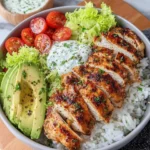 Close-up of a homemade grilled chicken rice bowl with sliced grilled chicken, fluffy white rice, avocado, cherry tomatoes, lettuce, and creamy herb sauce served in a white bowl on a marble kitchen counter with natural window light.