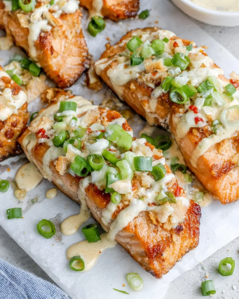 Close-up of homemade Bang Bang Salmon topped with sliced green onions and coated in a creamy spicy bang bang sauce, served on a plate in a bright home kitchen with natural lighting.