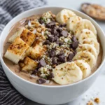 Chunky Monkey Smoothie Bowl topped with fresh banana slices, maple cinnamon granola, cacao nibs, hemp seeds, and chocolate chips in a white bowl on a marble countertop. Thick and creamy chocolate banana smoothie base with a healthy homemade breakfast presentation.