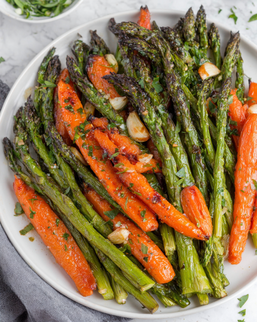 A white serving dish filled with roasted asparagus and carrots seasoned with garlic, olive oil, herbs, and black pepper. The vegetables are golden brown with caramelized edges and photographed in natural light on a white marble countertop in a cozy home kitchen.