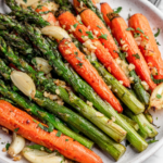 A white serving dish filled with roasted asparagus and carrots seasoned with garlic, olive oil, herbs, and black pepper. The vegetables are golden brown with caramelized edges and photographed in natural light on a white marble countertop in a cozy home kitchen.