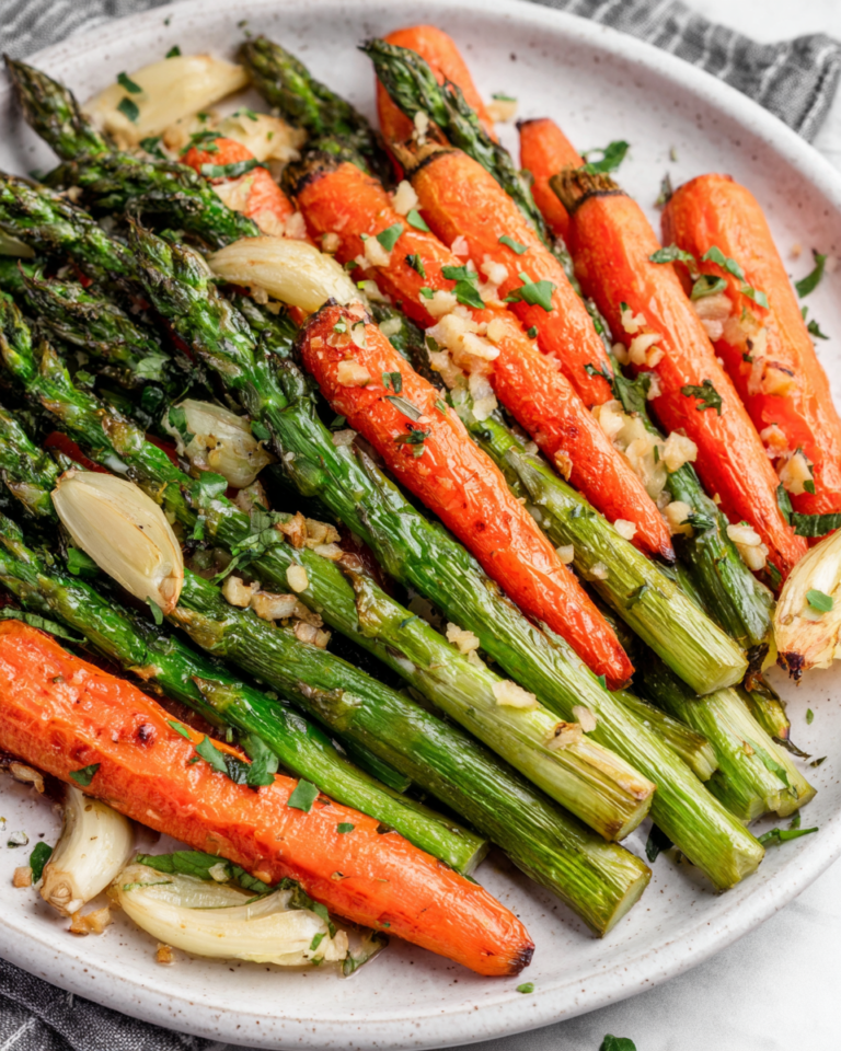 A white serving dish filled with roasted asparagus and carrots seasoned with garlic, olive oil, herbs, and black pepper. The vegetables are golden brown with caramelized edges and photographed in natural light on a white marble countertop in a cozy home kitchen.