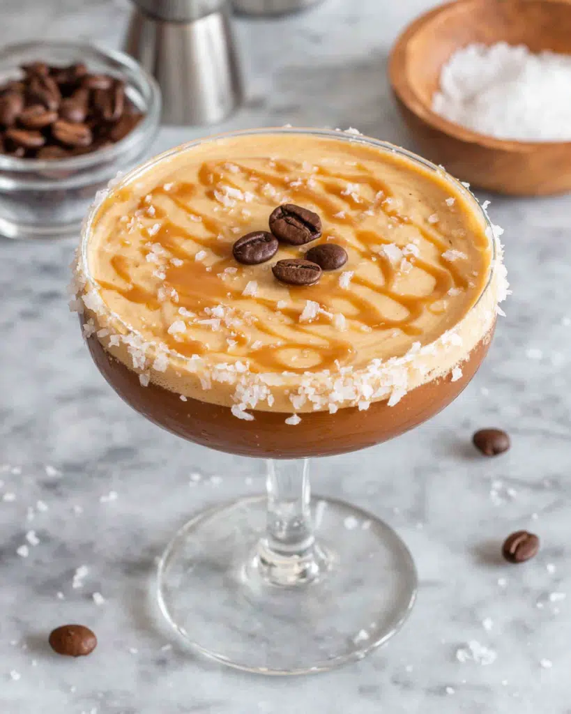 Salted Caramel Espresso Martini served in a coupe glass with a thick frothy espresso foam, three coffee beans on top, and a caramel sugar rim. The cocktail sits on a white marble countertop in a cozy home kitchen with natural window light and coffee beans scattered nearby.