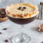 Salted Caramel Espresso Martini served in a coupe glass with a thick frothy espresso foam, three coffee beans on top, and a caramel sugar rim. The cocktail sits on a white marble countertop in a cozy home kitchen with natural window light and coffee beans scattered nearby.