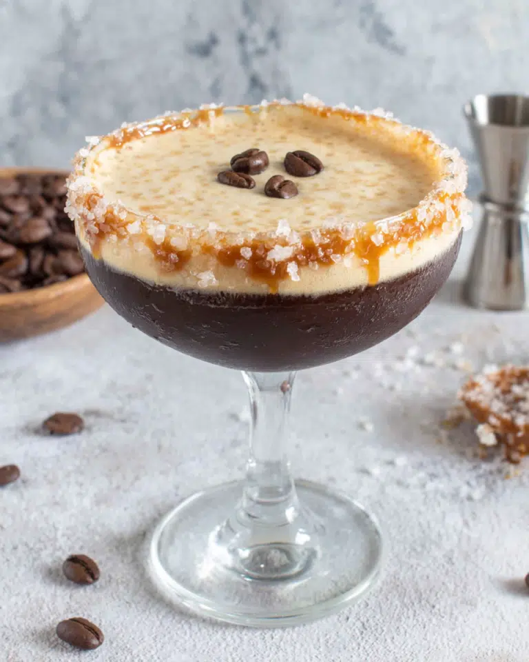 Salted Caramel Espresso Martini served in a coupe glass with a thick frothy espresso foam, three coffee beans on top, and a caramel sugar rim. The cocktail sits on a white marble countertop in a cozy home kitchen with natural window light and coffee beans scattered nearby.