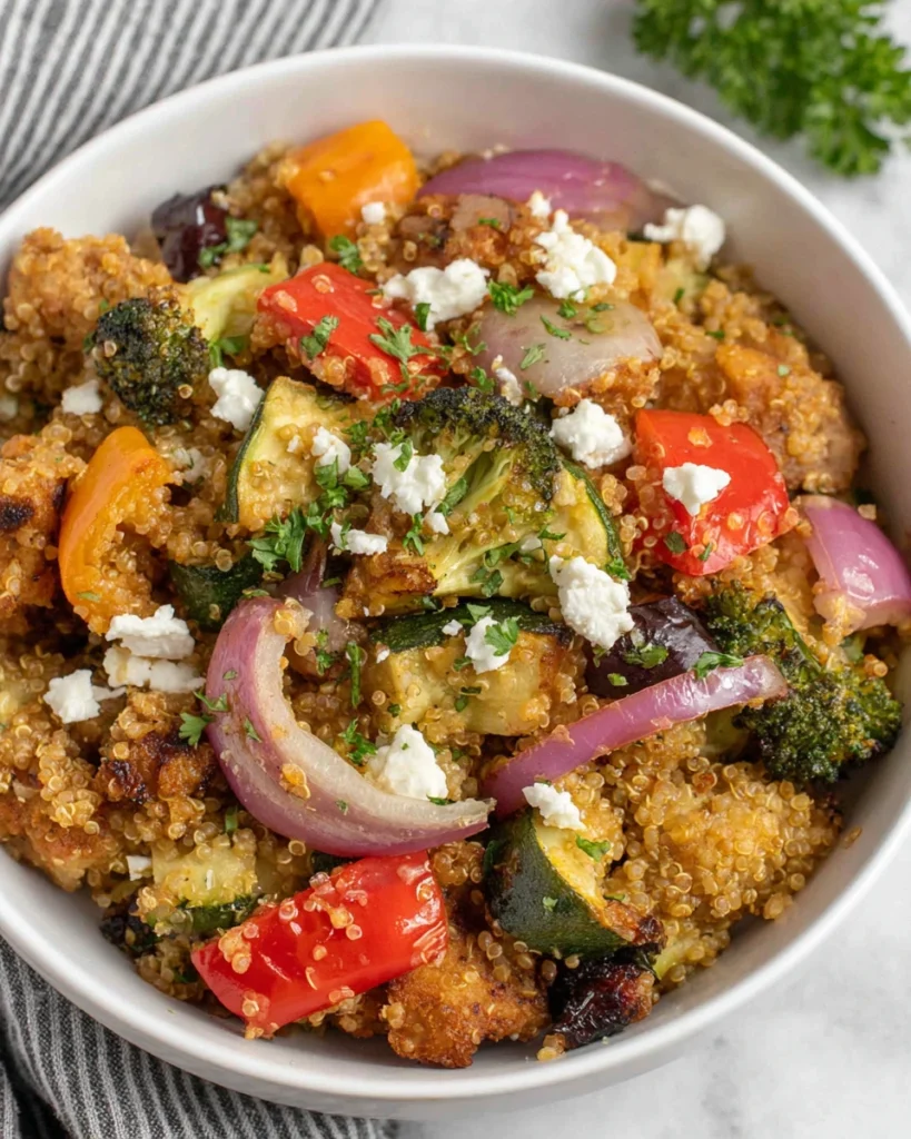 A bowl of Satisfying Sheet Pan Roasted Vegetable Quinoa Bake featuring fluffy quinoa, roasted bell peppers, zucchini, broccoli, cherry tomatoes, red onion, crumbled feta cheese, and fresh parsley. The dish is photographed in natural window light on a white marble countertop with a warm homemade kitchen atmosphere.
