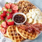 A homemade waffle board arranged on a wooden serving board with warm waffles, fresh strawberries, grapes, banana slices, peach wedges, crispy bacon, scrambled eggs, yogurt, walnuts, chocolate chips, and maple syrup, perfect for a family brunch or weekend breakfast.
