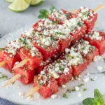 Watermelon Tajin Skewers arranged on a serving platter, topped with Tajín seasoning, fresh lime juice, crumbled cotija cheese, chopped mint, and flaky salt. A colorful sweet and spicy summer appetizer made with fresh watermelon and simple ingredients.