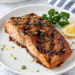 Close-up of zesty grilled honey mustard salmon topped with fresh parsley, dill, and lemon zest on a white plate. The salmon features a caramelized honey mustard glaze, beautiful grill marks, and a juicy flaky texture, photographed in natural window light on a white marble countertop.