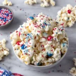 Close-up of homemade Patriotic Popcorn coated with red, white, and blue candy melts and topped with colorful M&Ms and festive star sprinkles. This easy Fourth of July snack is served on a tray and makes a fun party treat for summer celebrations.