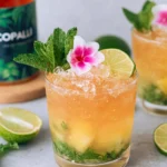 Mango Rum Swizzle cocktail served in a double old fashioned glass filled with crushed ice, made with white rum, mango juice, fresh lime juice, mint leaves, simple syrup, and bitters, garnished with a mint sprig, orchid flower, and lime round on a white marble countertop in natural light.