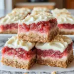 Freshly made Rhubarb Bars with Cream Cheese topped with a smooth cream cheese layer over sweet-tart rhubarb filling and a buttery oat crust, sliced into squares and served on a white plate in natural light.