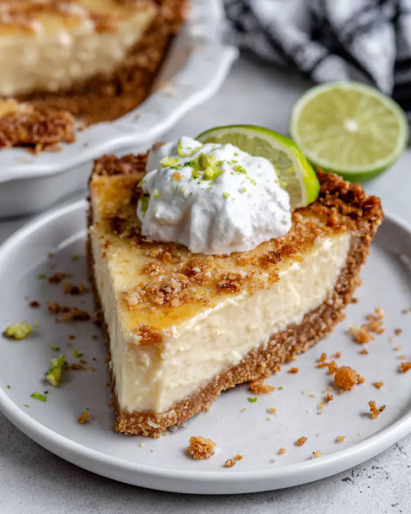 A slice of homemade 7-Ingredient Key Lime Pie with a buttery graham cracker and macadamia nut crust, creamy key lime filling, whipped cream, fresh lime zest, and lime slices on a white marble countertop in natural window light.