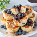 A plate of fluffy Easy 15-Minute Lemon Blueberry Cottage Cheese Pancake Bites topped with fresh blueberries and lemon zest, served on a white plate with maple syrup in natural window light, creating a cozy homemade breakfast scene.