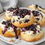 Close-up of homemade Gluten Free Blueberry Cheesecake Cookies with buttery lemon cookie dough swirled with blueberry jam, filled with creamy vanilla cheesecake, and topped with fresh blueberry sauce on a white marble countertop in natural window light.