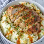 Juicy grilled chicken sliced over creamy Yukon Gold mashed potatoes in a white serving bowl, garnished with fresh chives and a drizzle of barbecue sauce. A comforting homemade dinner photographed in natural light on a white marble countertop.