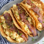 Savory Pancake Tacos filled with fluffy soft scrambled eggs, crispy bacon, melted cheddar cheese, and fresh chives inside golden homemade pancakes on a white plate, photographed in natural light with a cozy homemade breakfast presentation.