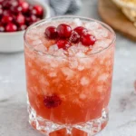A sparkling pink Sleeping Beauty Cocktail served over ice in a crystal rocks glass, garnished with maraschino cherries on a white marble countertop in a bright home kitchen.