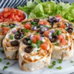 A plate of homemade Taco Cream Cheese Pinwheels made with cream cheese, cheddar cheese, taco seasoning, lettuce, tomatoes, black olives, and green onions wrapped in flour tortillas and sliced into bite-sized rolls on a white plate in a bright home kitchen.
