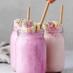 Creamy Pink Protein Smoothie made with frozen banana, Greek yogurt, vanilla protein powder, almond milk, and pink pitaya powder in a clear glass on a white marble countertop.