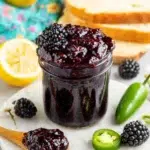 A glass mason jar filled with glossy homemade Blackberry Jalapeno Jam sits on a white marble countertop beside fresh blackberries, sliced jalapeños, lemon halves, and toasted artisan bread. The sweet and spicy jam has a rich deep-purple color with visible blackberry seeds, creating an inviting homemade presentation in natural kitchen lighting.
