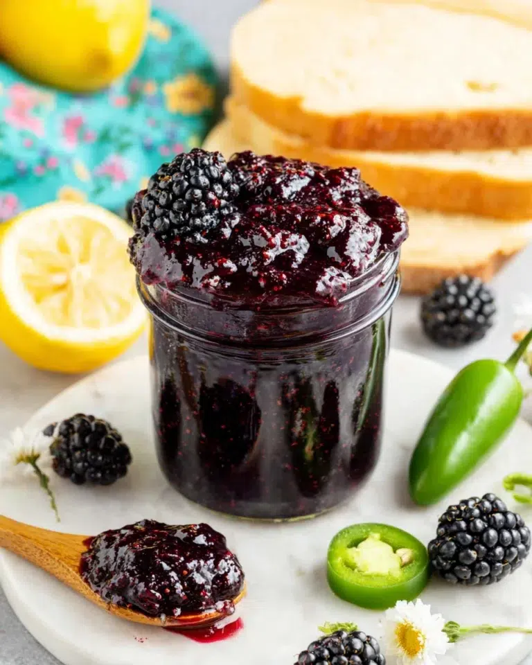 A glass mason jar filled with glossy homemade Blackberry Jalapeno Jam sits on a white marble countertop beside fresh blackberries, sliced jalapeños, lemon halves, and toasted artisan bread. The sweet and spicy jam has a rich deep-purple color with visible blackberry seeds, creating an inviting homemade presentation in natural kitchen lighting.
