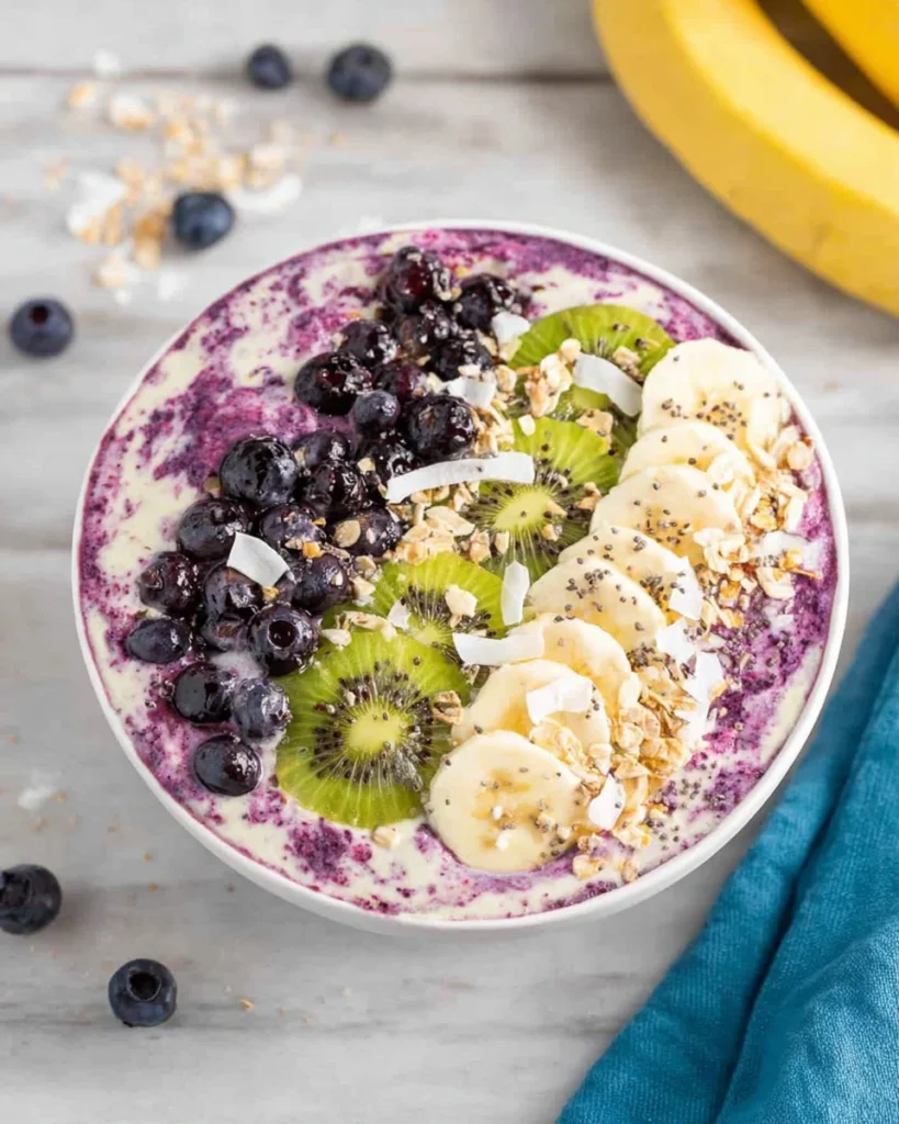 A thick and creamy Blueberry Banana Smoothie Bowl made with frozen blueberries, frozen banana, vanilla Greek yogurt, and milk, topped with fresh blueberries, banana slices, kiwi, coconut flakes, and chia seeds in a white bowl on a marble countertop.