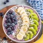 A thick and creamy Blueberry Banana Smoothie Bowl made with frozen blueberries, frozen banana, vanilla Greek yogurt, and milk, topped with fresh blueberries, banana slices, kiwi, coconut flakes, and chia seeds in a white bowl on a marble countertop.