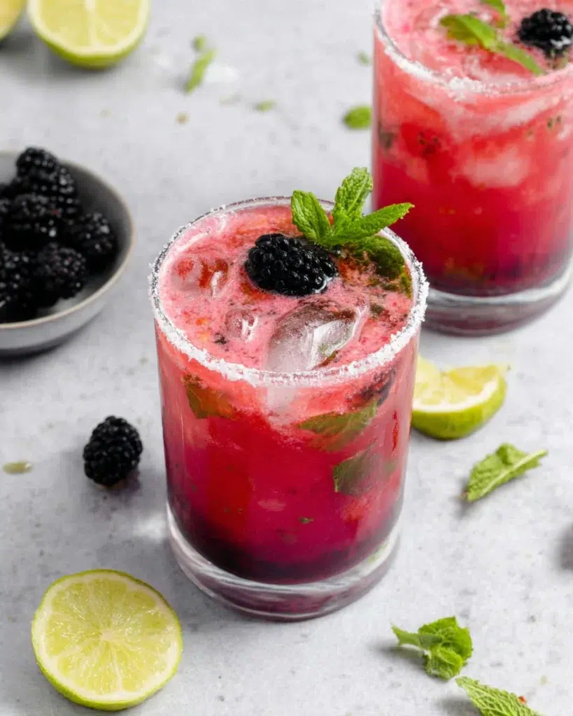 Creamy Blackberry Mojito Recipe served over ice in a sugar-lime rimmed glass, made with fresh blackberries, mint leaves, cream of coconut, lime juice, white rum, and sparkling soda water, garnished with a mint sprig, blackberry, and lime wheel on a white marble countertop in natural light.