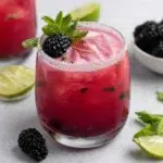 Creamy Blackberry Mojito Recipe served over ice in a sugar-lime rimmed glass, made with fresh blackberries, mint leaves, cream of coconut, lime juice, white rum, and sparkling soda water, garnished with a mint sprig, blackberry, and lime wheel on a white marble countertop in natural light.