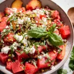 A colorful bowl of Watermelon Peach Salad made with juicy watermelon cubes, fresh peach slices, creamy crumbled feta cheese, basil leaves, roasted pistachios, and a homemade honey champagne vinaigrette, perfect for a refreshing summer meal.
