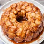 Freshly baked Granny's Monkey Bread Recipe served on a white plate, featuring golden pull-apart biscuit dough coated in buttery cinnamon sugar caramel. Homemade monkey bread photographed in natural kitchen light with a warm, gooey texture and rustic presentation, perfect for breakfast, brunch, or dessert.