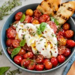 Close-up of Marinated Cherry Tomatoes with Burrata served on a white platter with juicy marinated cherry tomatoes, creamy torn burrata, fresh basil, thyme, cracked black pepper, and grilled bread on the side.