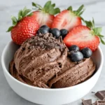 A bowl of creamy homemade chocolate frozen yogurt topped with dark chocolate shavings and fresh strawberries on a white marble countertop in a bright home kitchen.