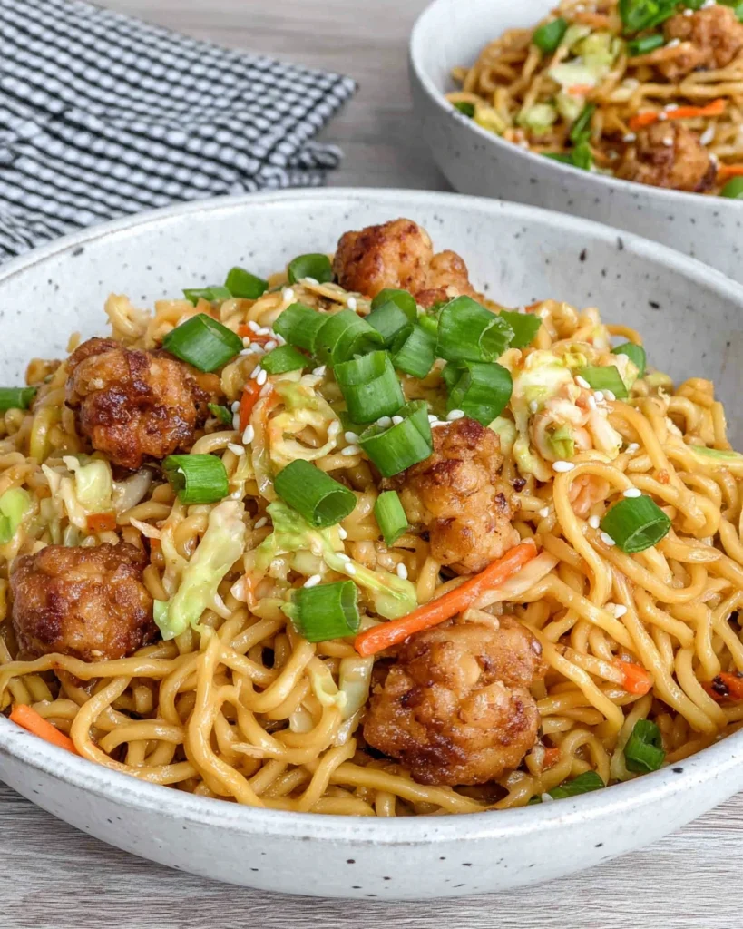 A bowl of Sticky Honey Garlic Chicken Noodles topped with crispy golden ground chicken, tender-crisp shredded vegetables, sliced scallions, and toasted sesame seeds, served on a white marble kitchen counter.