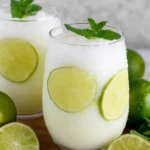 A glass of creamy Easy Brazilian Lemonade (Limeade) served over ice with fresh lime slices and mint on a white marble countertop, made with fresh limes, sweetened condensed milk, and cold water in a bright home kitchen.