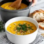 A bowl of creamy golden-orange butternut squash soup topped with fresh parsley and toasted pepitas, served with crusty bread on a white marble counter in natural window light.