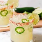 Spicy Margarita in a glass with a chipotle salt rim, fresh lime wedges, jalapeño slices, tequila, and ice on a white marble kitchen counter.