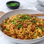 A bowl of Sticky Honey Garlic Chicken Noodles topped with crispy golden ground chicken, tender-crisp shredded vegetables, sliced scallions, and toasted sesame seeds, served on a white marble kitchen counter.