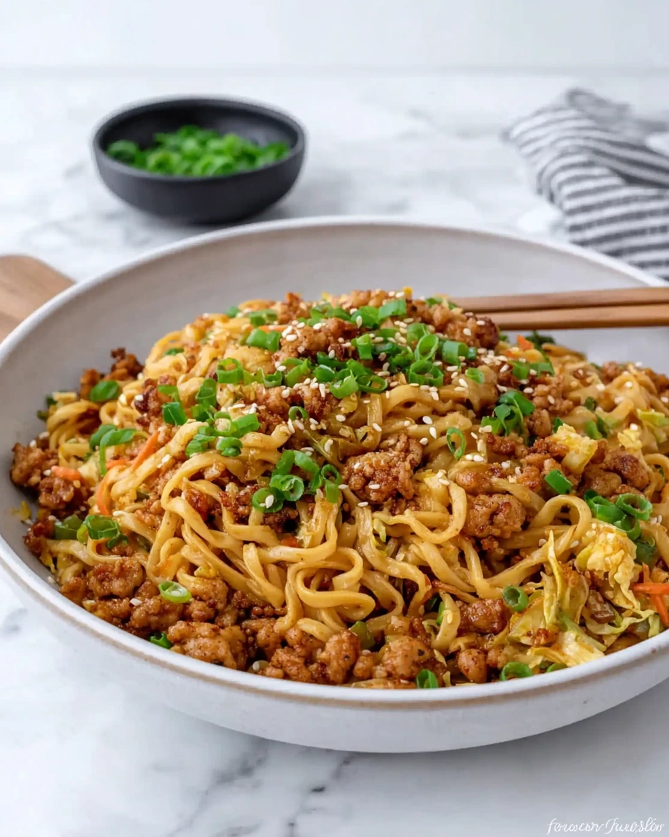 A bowl of Sticky Honey Garlic Chicken Noodles topped with crispy golden ground chicken, tender-crisp shredded vegetables, sliced scallions, and toasted sesame seeds, served on a white marble kitchen counter.