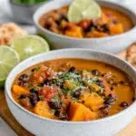 A bowl of sweet potato black bean soup with tender sweet potatoes, black beans, fire-roasted tomatoes, and fresh lime on a wooden board in a home kitchen.