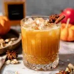 Spicy Bourbon Pumpkin Punch in a glass over ice, made with pumpkin puree, maple syrup, bourbon, orange juice, pumpkin pie spice, and optional ginger beer, photographed on a white marble kitchen counter.