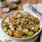 A bowl of Viral Green Crispy Potato Salad made with crispy roasted baby potatoes coated in a creamy green herb dressing, topped with fresh parsley, dill, crispy fried shallots, and toasted pumpkin seeds on a white marble countertop in natural window light.
